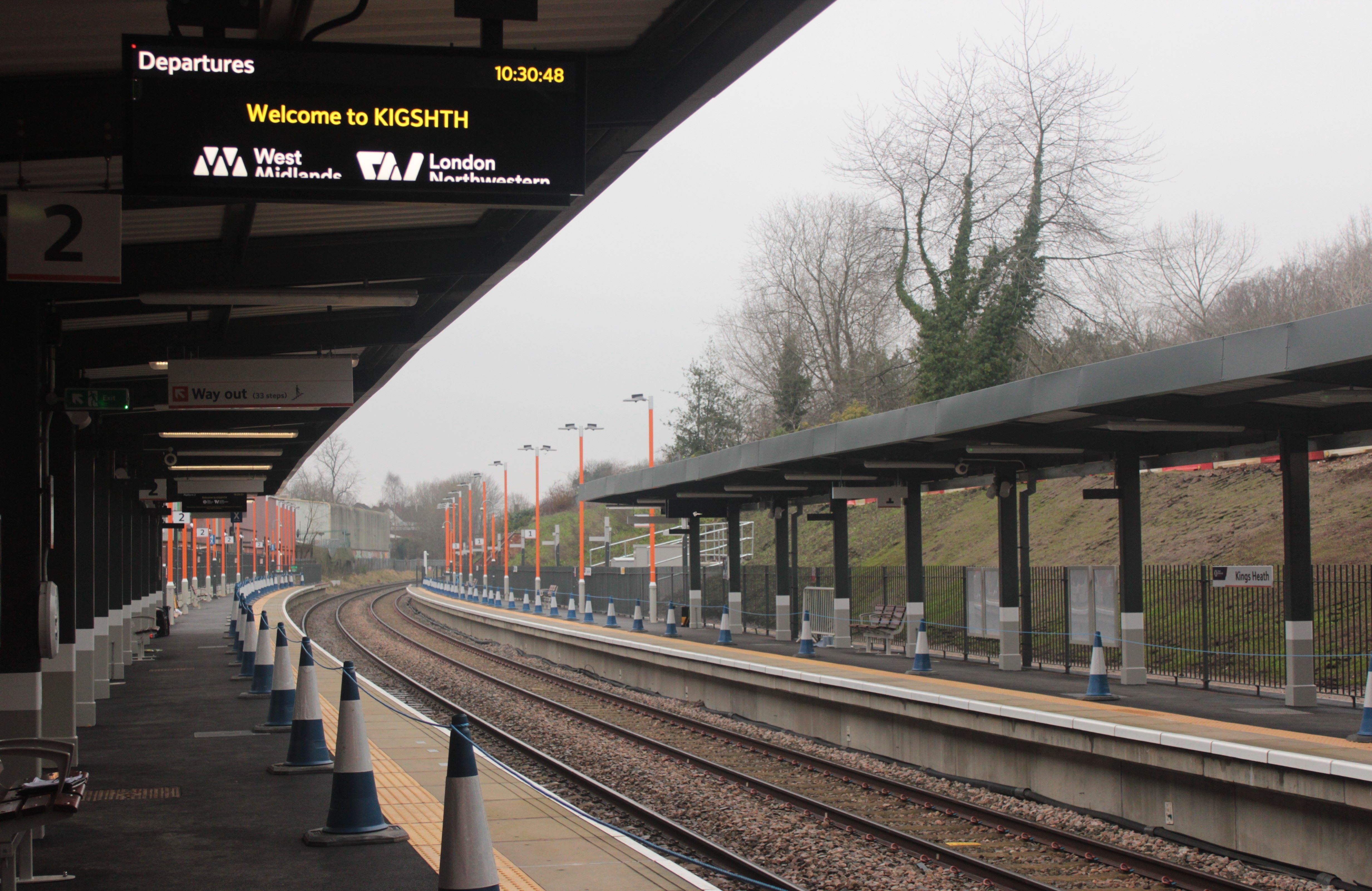 Railway station platform - tracks, platforms either side with canopies and sign in foreground saying welcome to Kings Heath